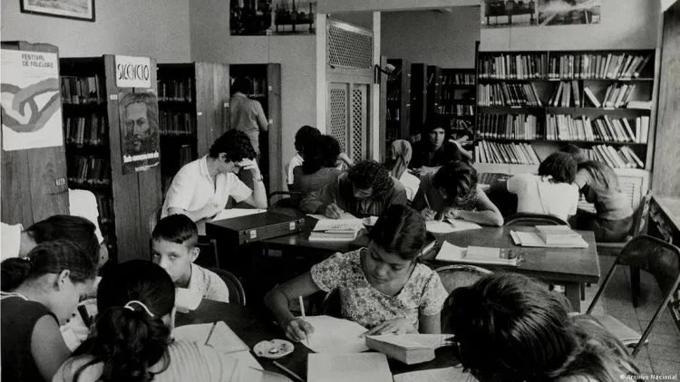 Foto em preto e branco mostra crianças sentadas ao redor de mesas em uma biblioteca